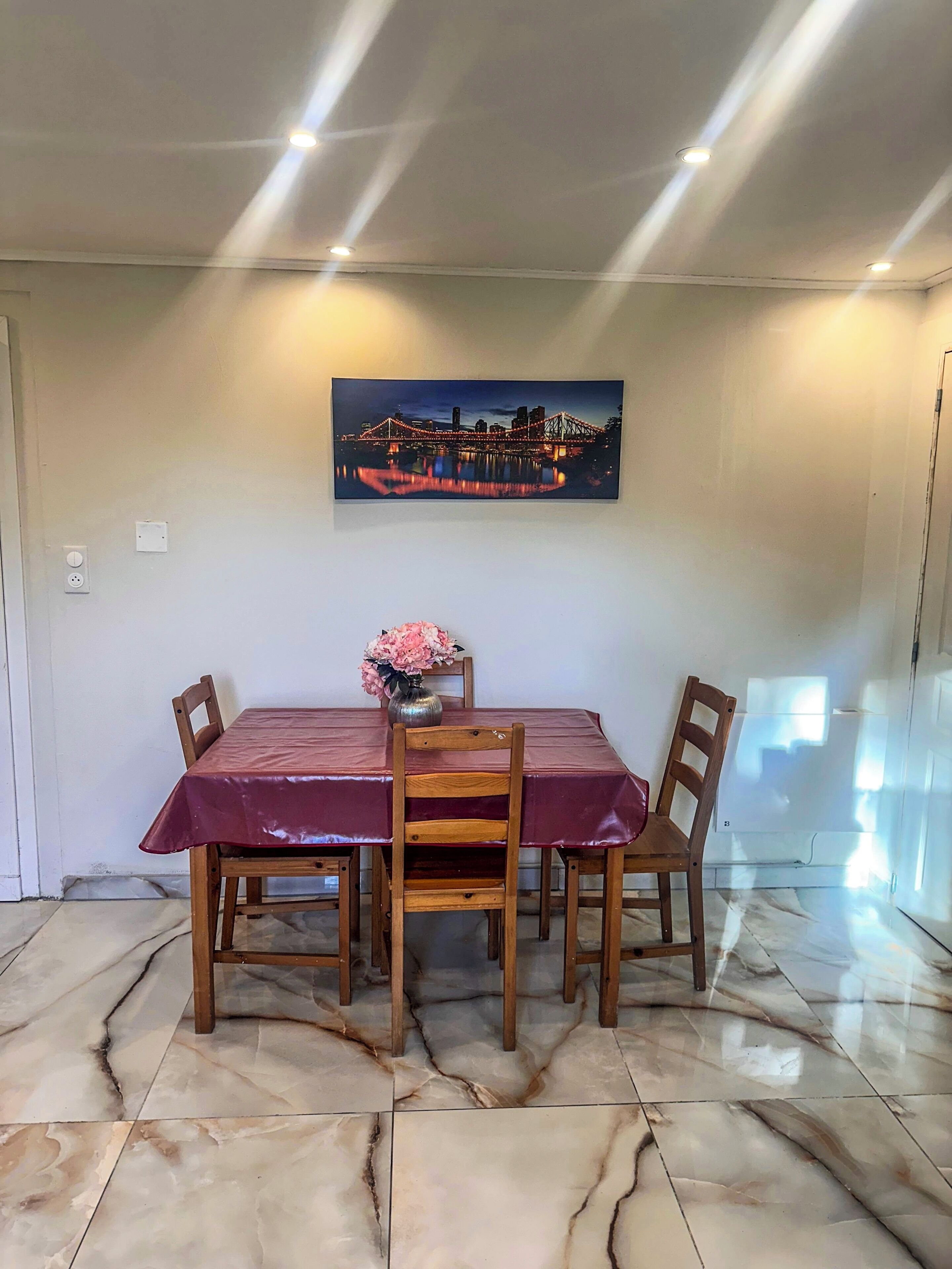 Un comedor con una mesa dispuesta para cuatro, un florero con flores y un cuadro de la ciudad en la pared.