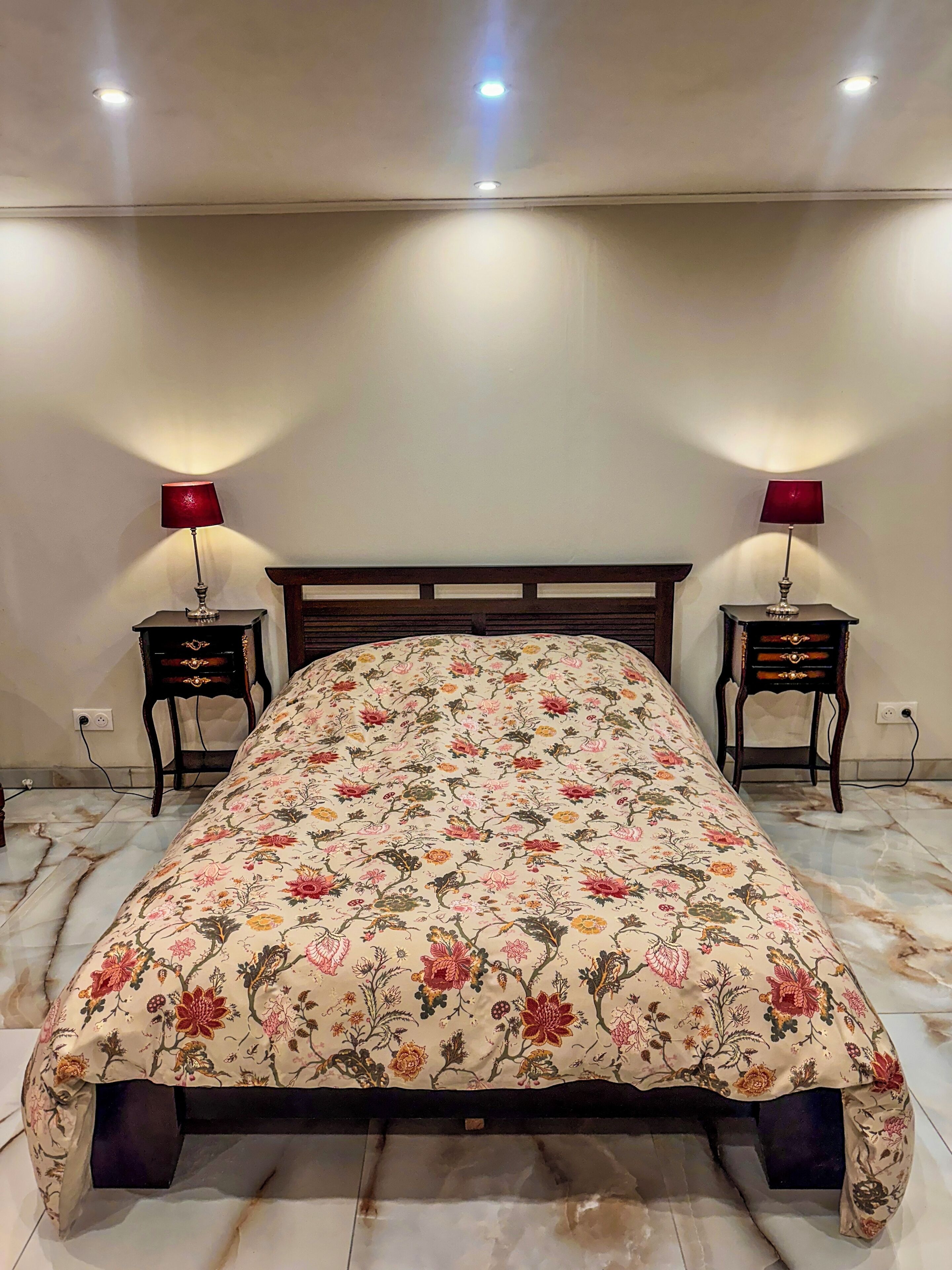 Un lit recouvert d’une couvre-lit à motifs floraux, deux tables de chevet avec des lampes et un sol en marbre.