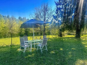 Ein Tisch und Stühle stehen unter einer Markise in einer grünen, von Bäumen gesäumten Außenanlage.