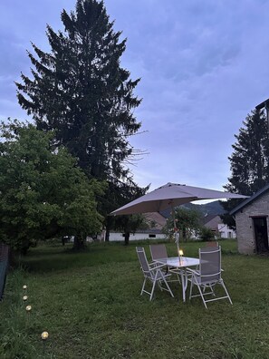 Eine Terrasse mit Tisch und Stühlen, umgeben von Gras und Bäumen, mit einem Gebäude im Hintergrund.