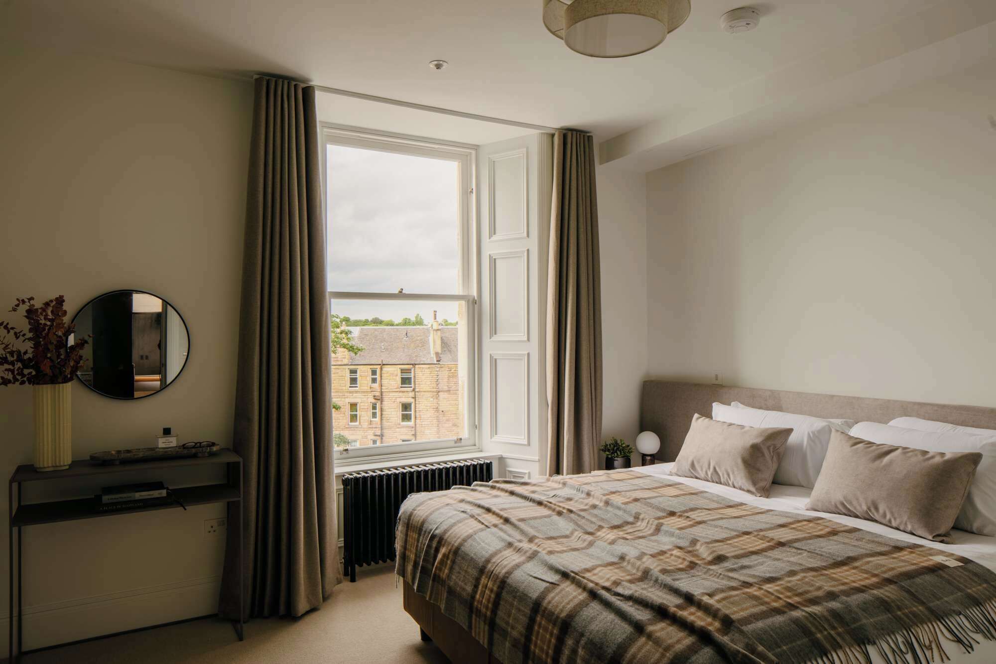 A bedroom with a bed, a bedside table, a radiator, a window with curtains, and a mirror on the wall.