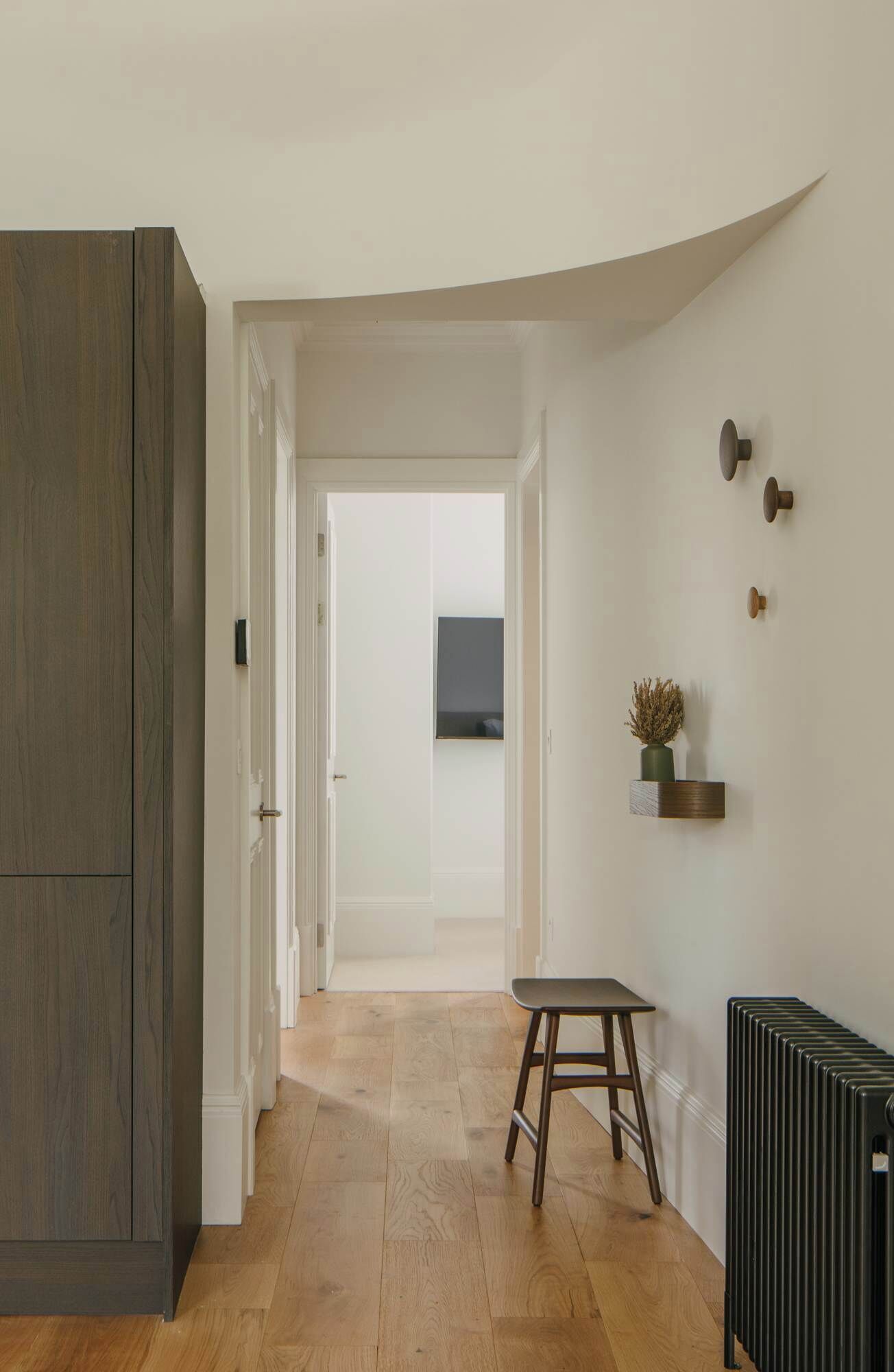 A hallway with a wooden wardrobe, a radiator, a small stool, and a wall-mounted shelf with a plant.
