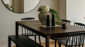 Et spiseområde med et rundt spejl, et træbord, sorte stole og en vase med hvide blomster.