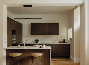 A modern kitchen with dark wood cabinets, a white countertop, and two bar stools. - Charming Apartment in Claremont Residences (Edinburgh)