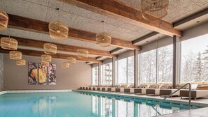 A swimming pool with wooden beams and hanging wicker lights, large windows with a view of trees, and a row of cushioned seating along the poolside. - Rothornblick 39 by Arosa Holiday Appartement/Fewo,   4-Bettwohnung by Interhome (Arosa)