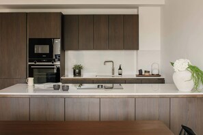 A modern kitchen with dark wood cabinets, a white countertop, a built-in oven, a microwave, a sink with a modern faucet, and a vase with flowers. - Stylish Apartment in Claremont Residences (Edinburgh)