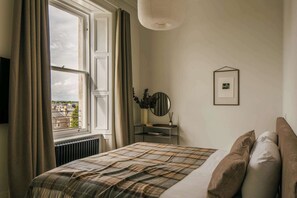A bedroom with a bed, a window with curtains, a radiator, a bedside table with a mirror, and a framed picture on the wall. - Refined Apartment in Claremont Residences (Edinburgh)