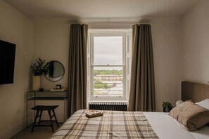 Ein Schlafzimmer mit Bett, Schreibtisch, Stuhl, einem Fenster mit Vorhängen und Blick auf Gebäude.