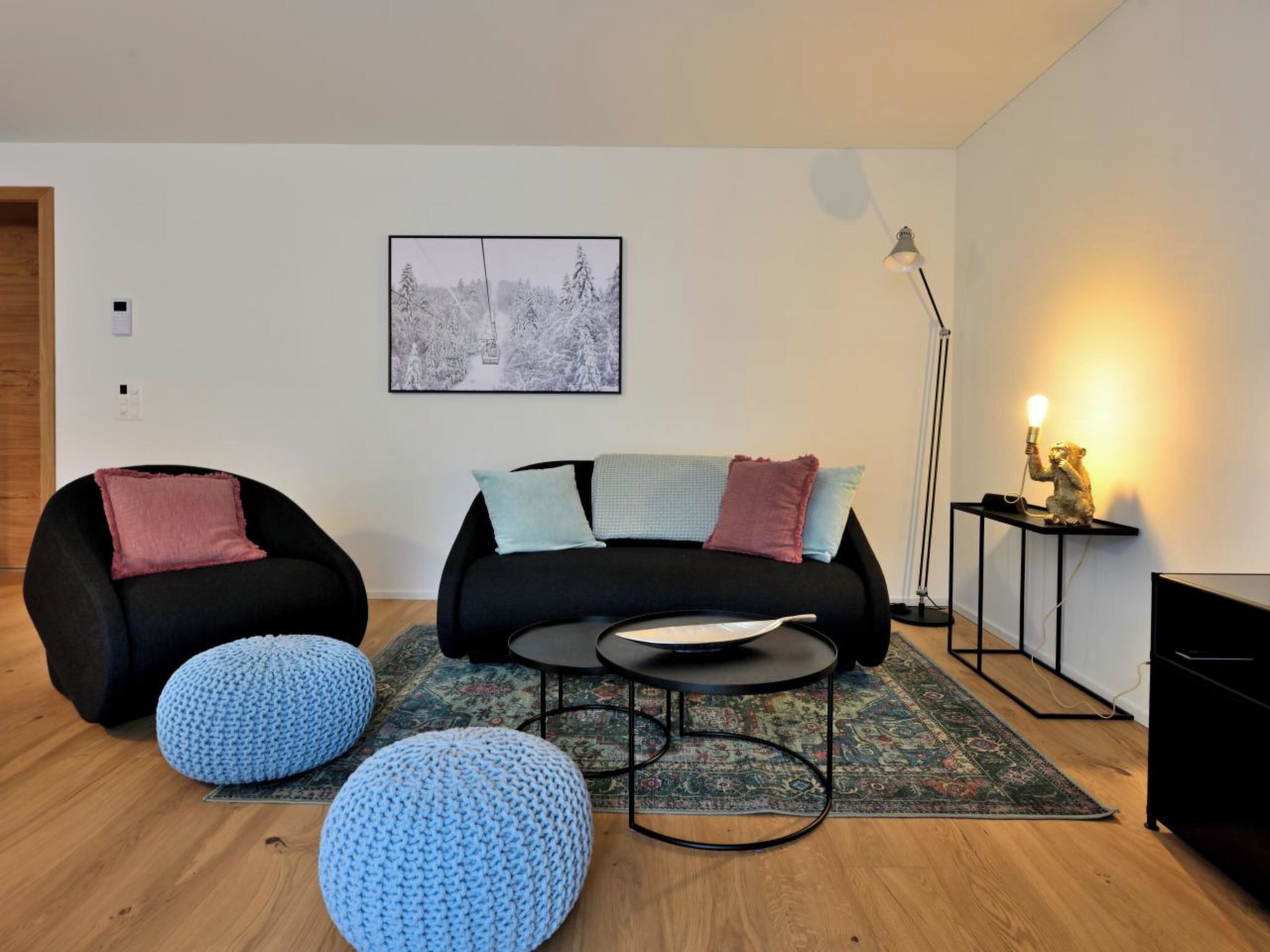 Un salon moderne avec un canapé noir, deux poufs bleus, une table basse ronde, un tableau encadré au mur, une lampe sur pied et une petite table avec une lampe.