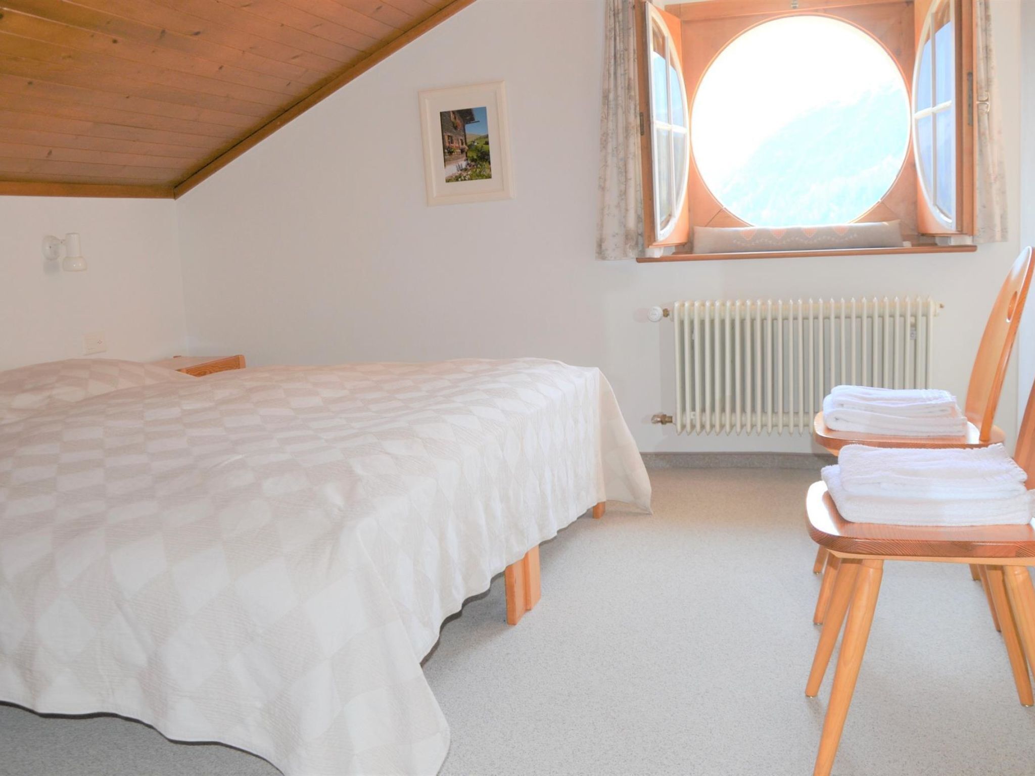 A bedroom with a bed, a chair with towels, a radiator, and a window with a view.