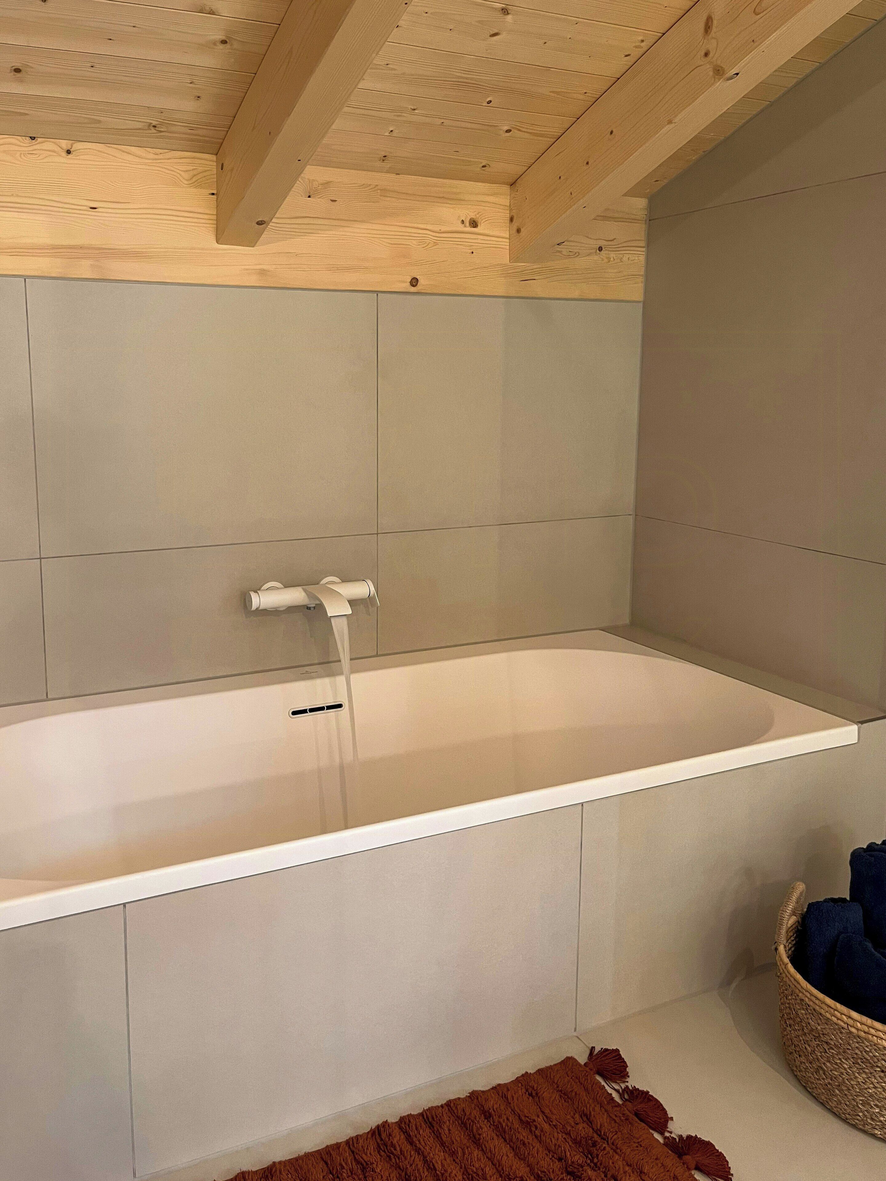 Un baño moderno con una bañera grande, vigas de madera en el techo y una paleta de colores neutros.