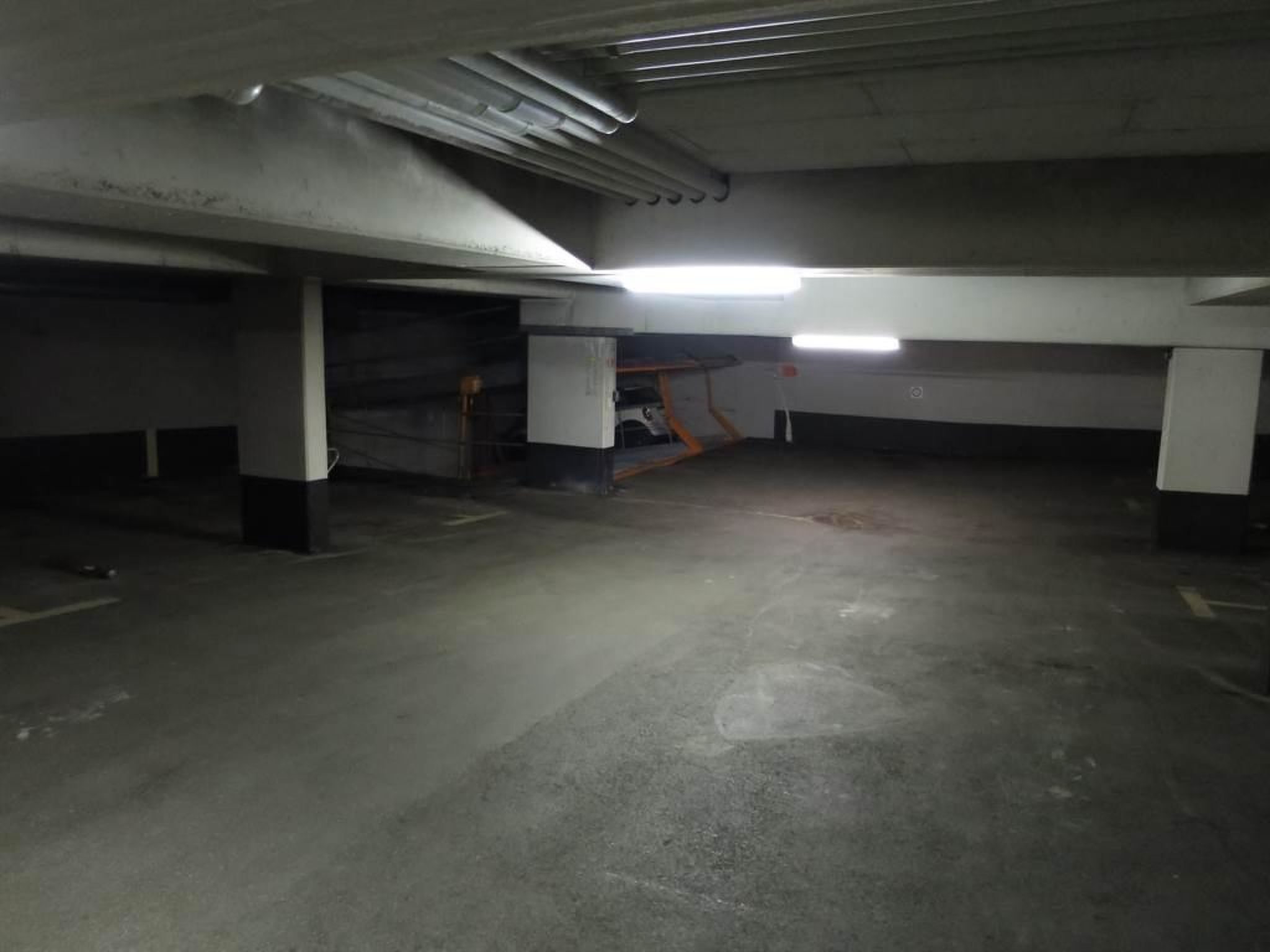 Un parking souterrain avec des piliers en béton, un éclairage fluorescent et une rampe d’accès clairement visible.