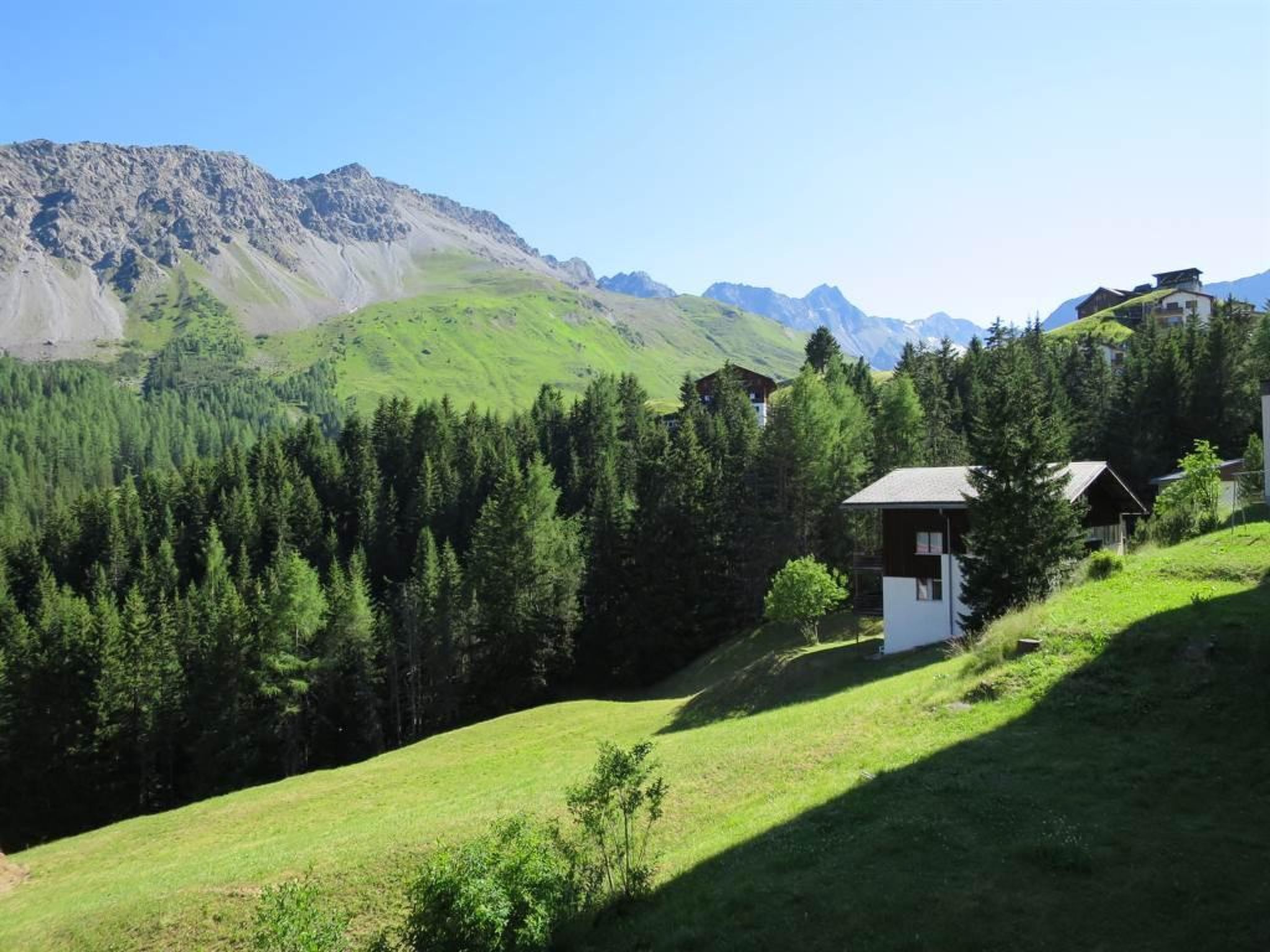 Eine gebirgige Landschaft mit einem kleinen Gebäude, umgeben von dichtem Wald und blauem Himmel.