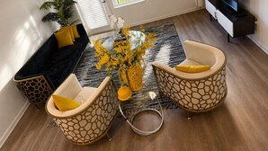 Ein Wohnzimmer mit einer Couch, zwei Sesseln, einem Couchtisch mit Blumen, einem Fernseher und einer Pflanze.