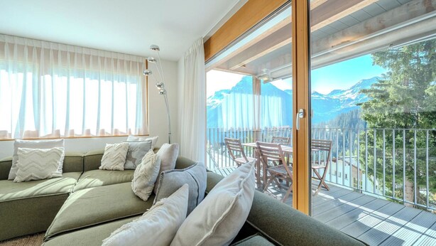 A modern living room with a green sofa, white cushions, and a large sliding glass door leading to a balcony with a mountain view. - Sunnaschy Heidi by Arosa Vacations Appartement/Fewo, Dusche und Badewanne, 2 Sch by Interhome (Arosa)