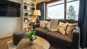 Uma sala de estar com sofá, apoio para os pés, mesa de centro e televisão na parede. Há uma estante com diversos objetos e uma janela com vista para o exterior.