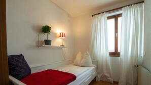 Quarto pequeno com uma cama de solteiro, piso de madeira, mesa de cabeceira com luminária, planta em vaso e janela com cortinas.