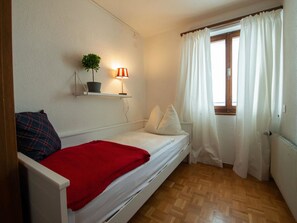 Ein kleines Zimmer mit einem Einzelbett, Holzboden, einem Nachttisch mit Lampe, einem Topfpflanzen und einem Fenster mit Vorhängen.