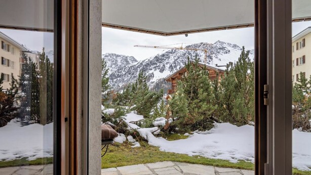 On aperçoit, à travers une porte vitrée, un patio en pierre qui mène à un paysage montagneux, avec des arbres couverts de neige et un chalet en bois.