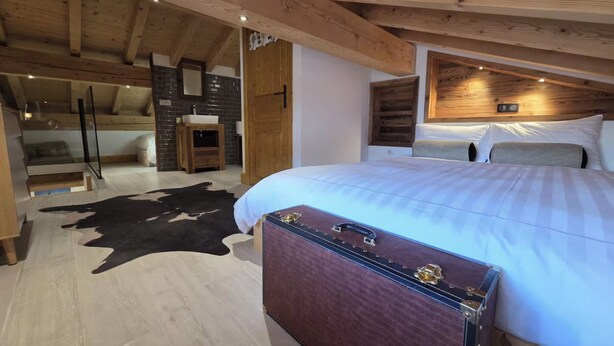Una camera da letto con soffitto in legno, un letto con biancheria bianca, un tappeto in pelle di vacca e una valigia vintage.