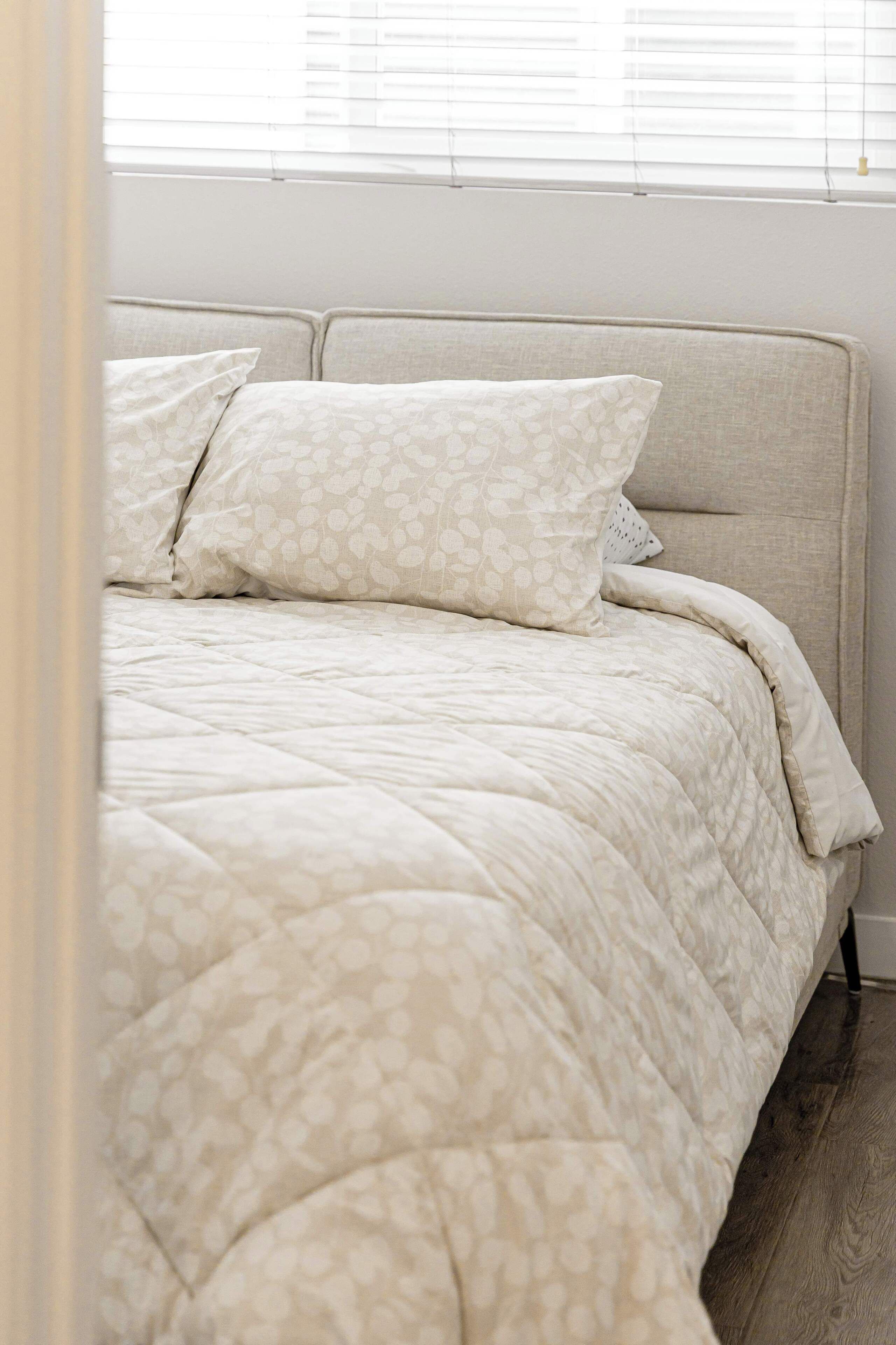 A bed with a patterned bedspread and pillows, a headboard, and a window with blinds.