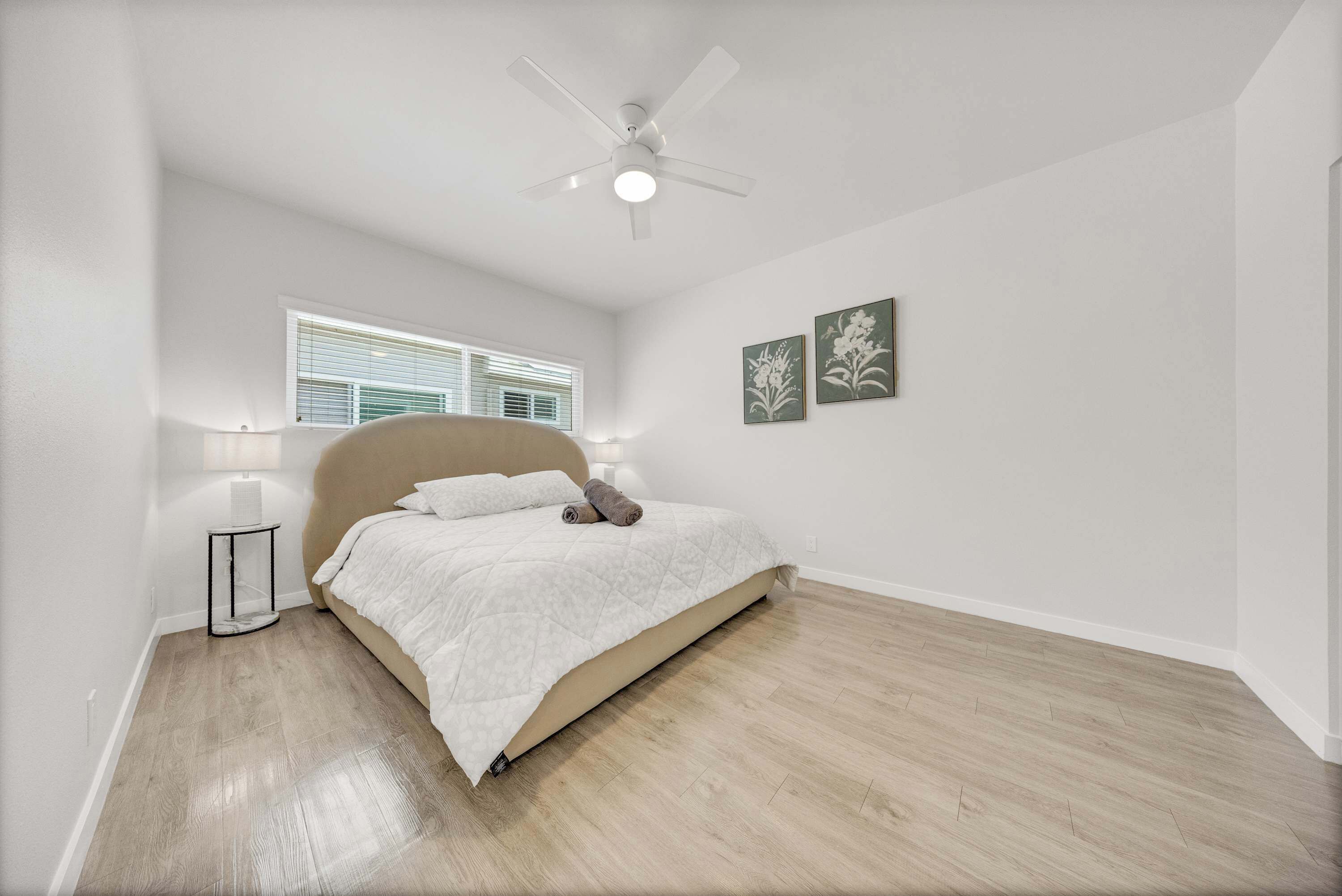 Un dormitorio con una cama, un ventilador de techo, una ventana con persianas y dos cuadros colgados en la pared.
