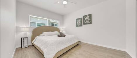 Un dormitorio con una cama, un ventilador de techo, una ventana con persianas y dos cuadros colgados en la pared.