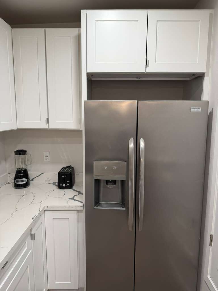 A stainless steel refrigerator with a water dispenser, a blender, and a toaster on a marble countertop in a kitchen with white cabinets.