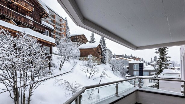 Schneebedeckte Außenanlage mit einem Holzbauten, einer Glasuhr und Blick auf weitere Gebäude.