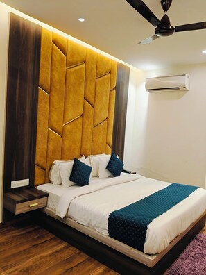 A bedroom with a tufted headboard, a bed with white and teal bedding, a wooden nightstand, a ceiling fan, and an air conditioning unit. - Ganges Blossom Hotel (Rishikesh)