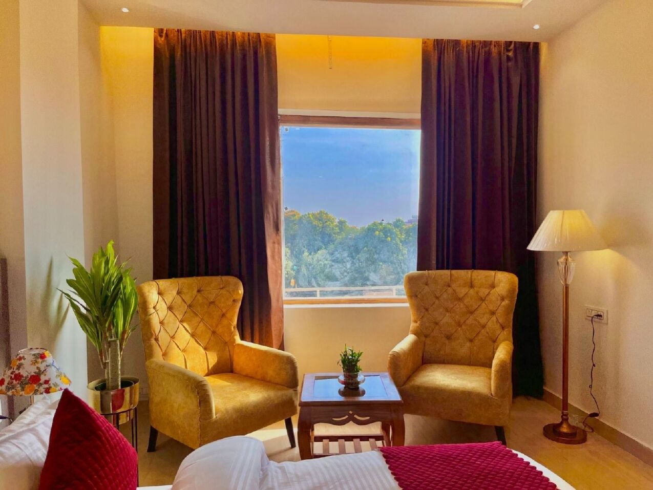 Habitació d'hotel amb dues cadires tapissades, una taula petita amb una planta, un llit amb llençol vermell i una finestra amb cortines.