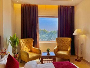 A hotel room with two upholstered chairs, a small table with a plant, a bed with a red blanket, and a window with curtains. - Ganges Blossom Hotel (Rishikesh)