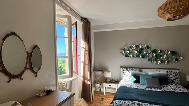 A bedroom with a large bed, a dresser, a bedside table, a lamp, and a view of the outdoors.