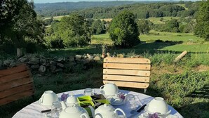 A round table set with white teacups, teapots, and a pitcher, surrounded by greenery and a stone wall.