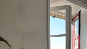 A room with a wooden dresser, a mirror, a window with a red frame, and a view of greenery and mountains.