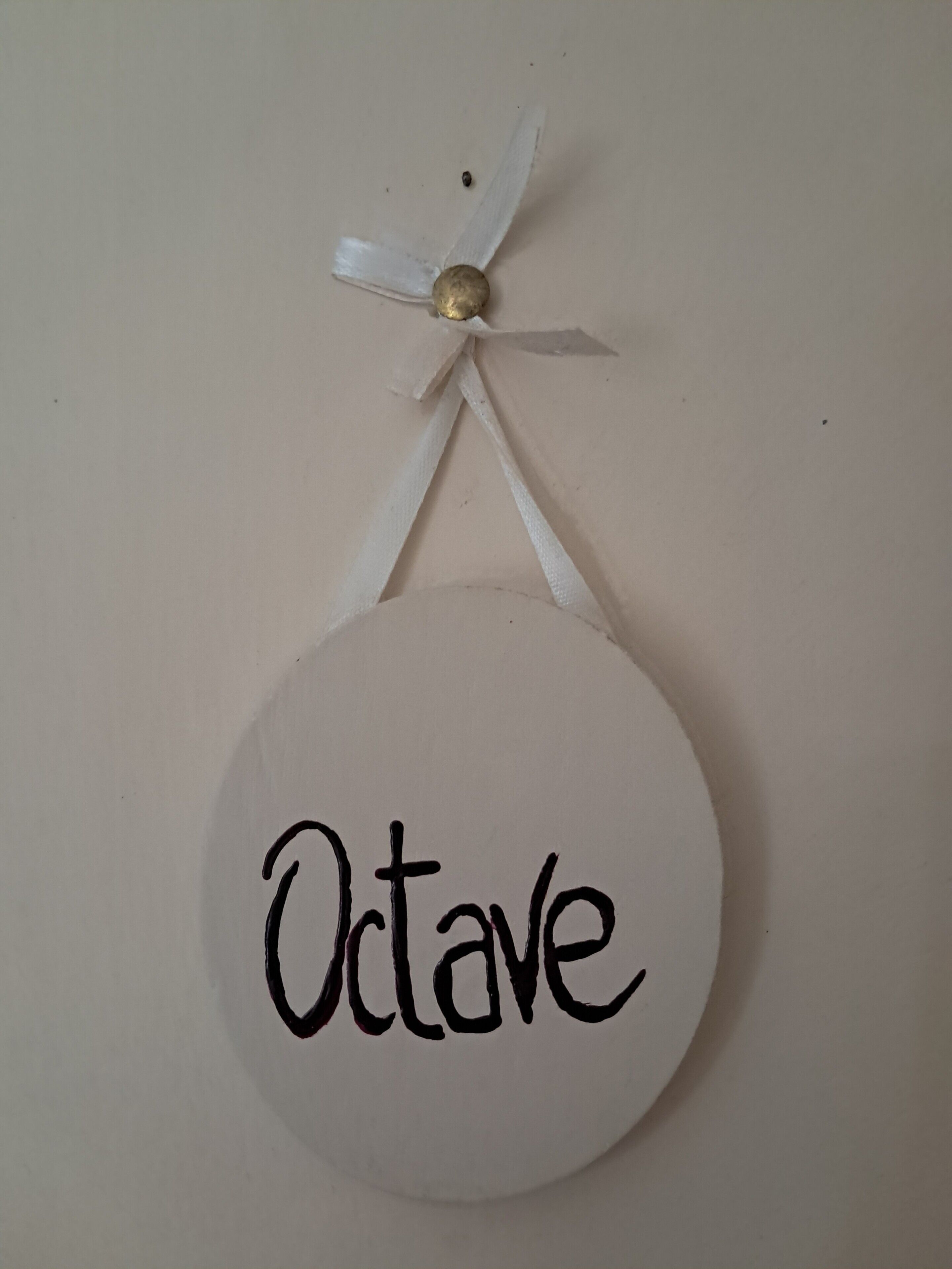 A round white sign with the word 'Octave' written on it, hanging from a white ribbon tied around a small golden bead.