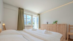 Un dormitorio con dos camas, un aparador de madera, una lámpara de noche y una ventana con vistas a montañas.