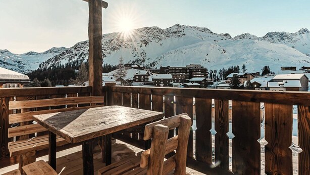 Een houten terras met banken en een tafel, met uitzicht op een met sneeuw bedekte berg en een dorp.