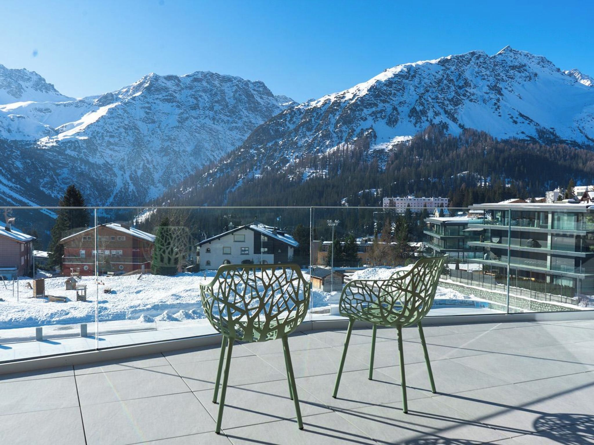 Un balcon avec deux chaises vertes, un paysage de montagnes enneigées en arrière-plan et des bâtiments visibles au loin.