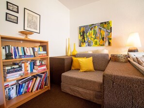 Ein gemütliches Wohnzimmer mit Bücherregal, einer gelben Kissensofa, einer Lampe und einem Bild an der Wand.