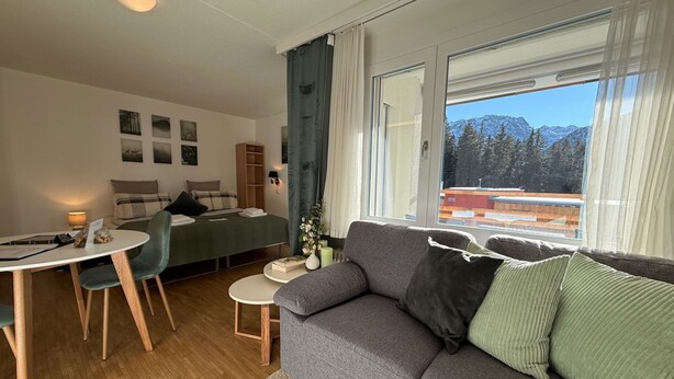 Een kamer met een bed, een slaapbank, een eettafel met stoelen en een raam met uitzicht op bergen.