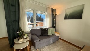 Een knus interieur met een grijze bank, een klein rond tafeltje met een vaas, en uitzicht op bergen door het raam.