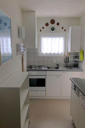 Eine kleine Küche mit weißen Schränken, einem Herd, einem Backofen, einer Spüle und einem Fenster mit Vorhängen.