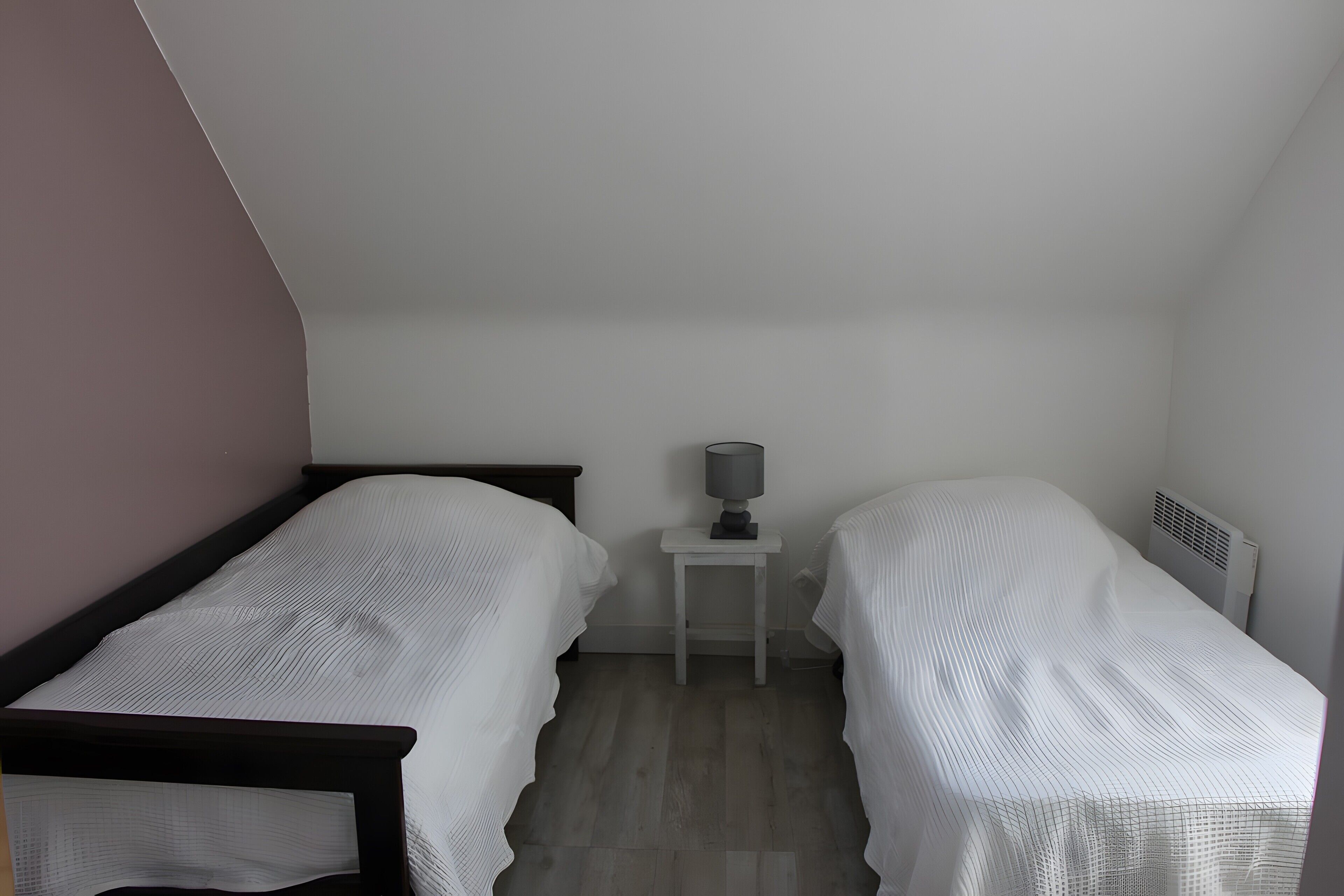 Una habitación pequeña con poca luz, con dos camas individuales, una mesita de noche con lámpara y un radiador.