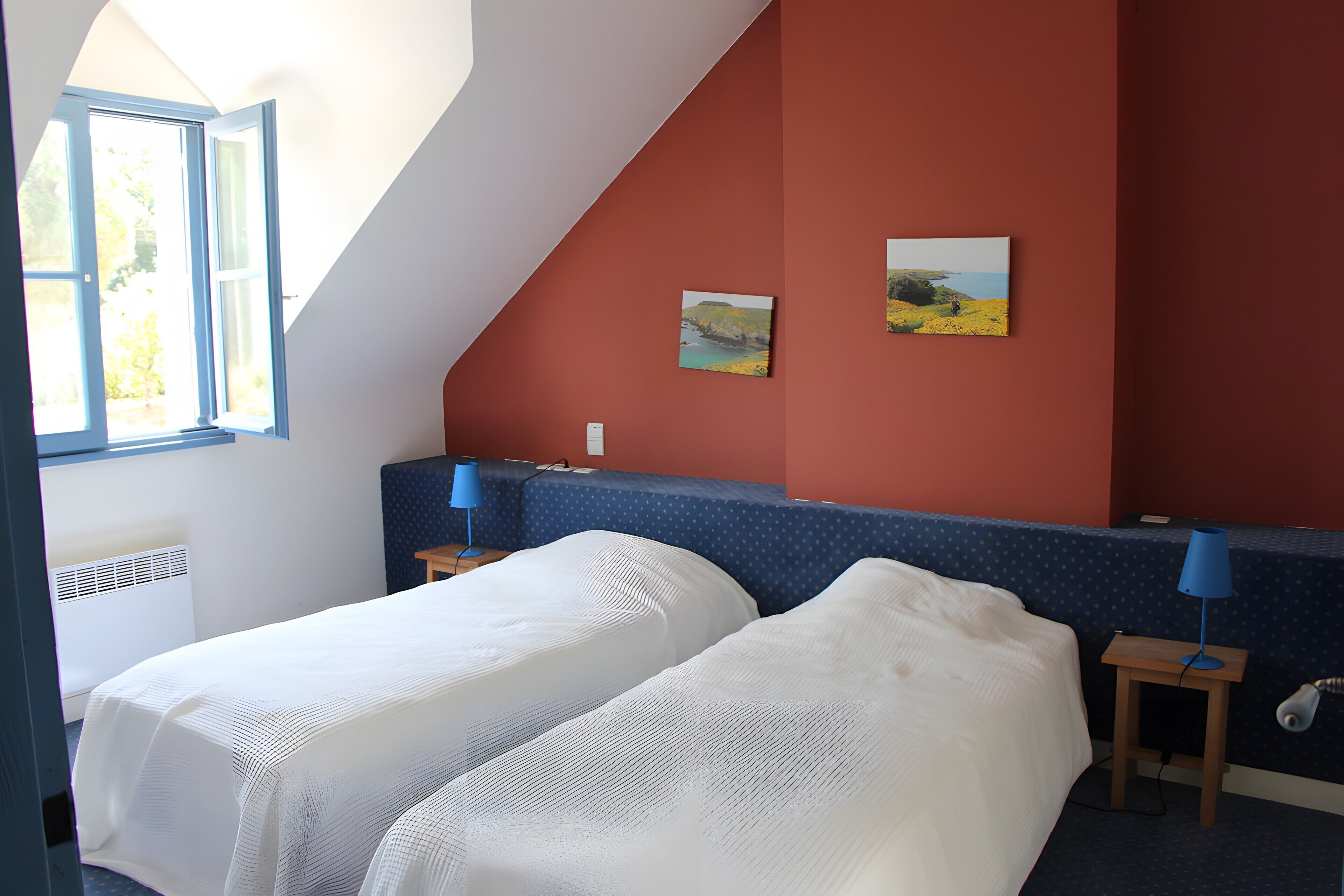 Une chambre avec deux lits, une fenêtre, un mur rouge et deux tableaux.