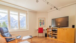 Ein Wohnzimmer mit einem großen Flachbildfernseher, einer Holzkassette mit Büchern, einem roten Sessel und einem Tisch.
