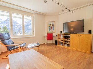 A living room with a large flat-screen TV, a wooden cabinet with books, a red chair, and a table. - Camana Wohnung EG Appartement/Fewo, Dusche oder Bad, WC, 6-Bettwohnung Camana EG by Interhome (Arosa)