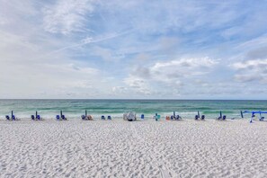 Ein Strand mit blauen Liegestühlen und Sonnenschirmen, feinem Sand und einem ruhigen Meer unter einem teils bewölkten Himmel.