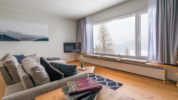 Een moderne woonkamer met een bank, een bijzettafeltje met boeken, een televisie en een groot raam met gordijnen.