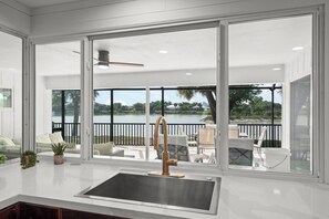 A modern kitchen with a large window, a stainless steel sink, and a view of a lake and outdoor seating area. - Murex Home – Luxury Sanibel Island Pool Home | Lake Views | Walk to Beach (Sanibel)